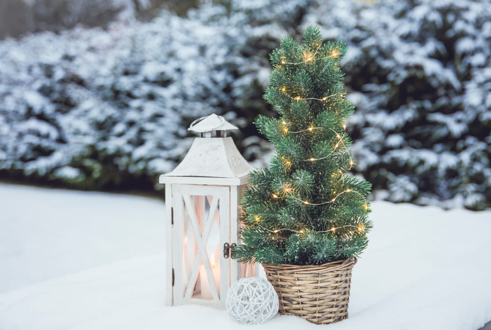 White wooden lantern with white wax candle lit and small Christmas fir tree with micro led lights in brown rattan flower pot in snow, snowy fir trees in the background, outdoors in winter
